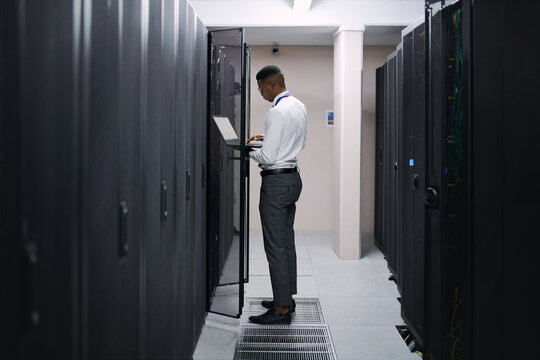 Engineer, black man or typing on laptop in server room for console access, power backup or update. Computer technician, person and inspection in data center for system upgrade, troubleshoot or review