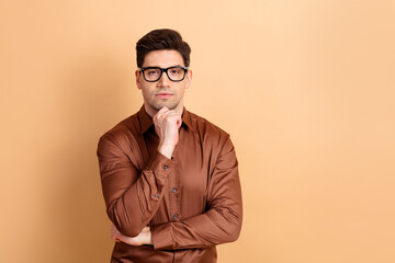 Obraz premium Thoughtful young man wearing glasses and brown shirt posing in a studio portrait with hand on chin, confident professional look and neutral beige background