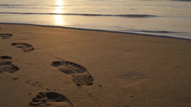 Human footprints on sandy beach being washed away by wave at sunset