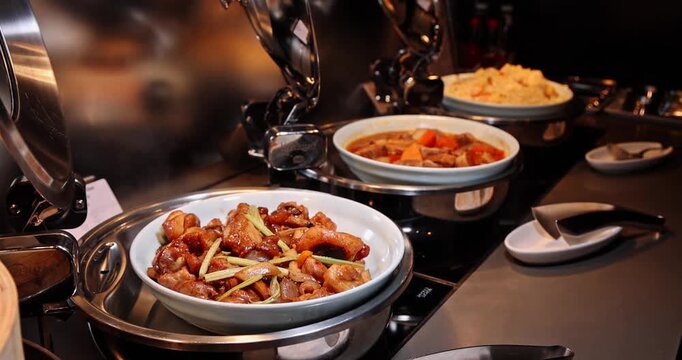 Assorted hot dishes including stir-fried chicken, meat stew with carrots, and fried rice served in stainless steel chafing dishes at a hotel buffet during dinner. Close-up side view. Slow motion.