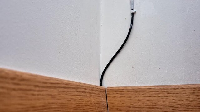 Room corner with intersecting white walls and wooden baseboard, showing black cable emerging from wall fitting, highlighting household wiring detail and simple construction element