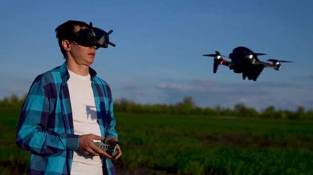 Flying drone with fpv goggles pilot wearing blue plaid shirt holds controller while quadcopter hovers over green field under clear sky for hobby flight and outdoor practice fpv training control