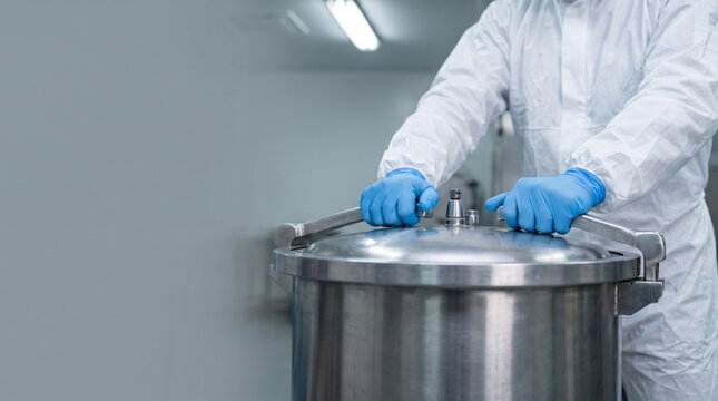 White protective suit worker sealing stainless steel autoclave drum lid with blue gloves, clinical waste sterilization, safety compliance training.
