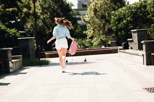 Young woman in casual summer outfit runs through a sunny park in the city carrying a pink backpack capturing a joyful outdoor moment of urban life and carefree style
