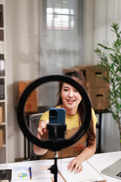 Asian female entrepreneur streaming live video with smartphone and ring light, creating content for social media marketing as a small business owner blogger