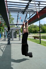 Naklejka premium A strong man in a black tracksuit hangs on special suspended hooks on the street sports ground. Modern workout area. 