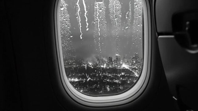 Rain drops stream down airplane window with nighttime city lights in black and white during flight