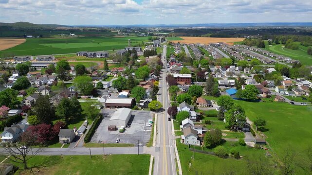 High-angle drone shot capturing a two-lane highway passing through lush agricultural fields and leading into a small residential community during a clear day.