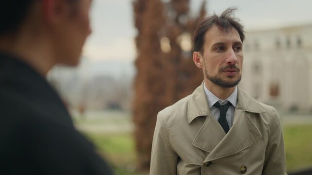Two business professionals engage in an important outdoor meeting, conversing in an urban setting. A man in a trench coat talks seriously with his colleague.