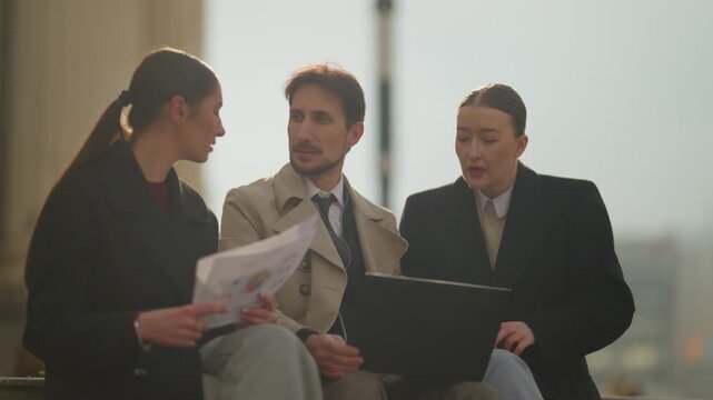 Three business people actively engage in a collaborative discussion with a laptop and documents. They are meeting outdoors, working on a project in a professional urban setting.