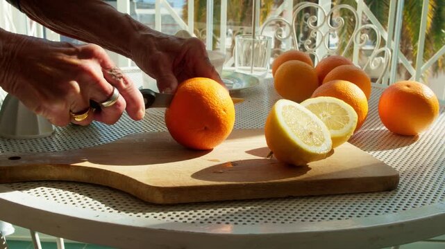 Close-up of hands cutting and juicing fresh oranges on a sunny outdoor terrace. Preparing a healthy natural breakfast with organic citrus fruits on a wooden board during a bright morning