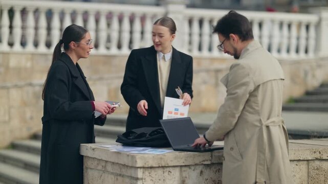 Business professionals collaborate on a project in an urban outdoor setting. They are reviewing documents and using digital devices for their work.