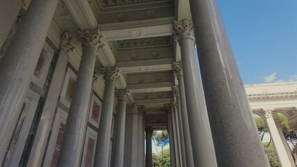 Naklejka premium Row of tall stone columns with ornate capitals supporting decorated ceiling in classical architecture, exterior view with blue sky and trees in the background