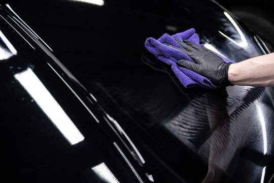 Close up of a detailer wiping off polishing compound from a black car surface after machine polishing. The step reveals a clean glossy finish and removes residue left from the correction process