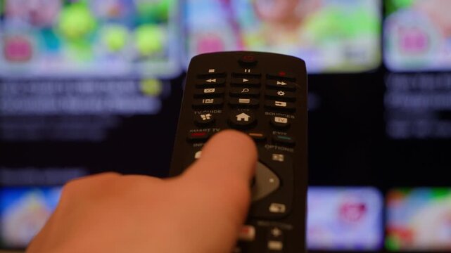 A close-up of a hand holding a TV remote control and pointing it at the screen. A blurred TV interface with streaming content appears in the background. Conceptually, this refers to home entertainment