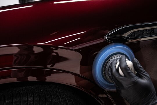 Close up of a professional detailer polishing red car paint using a machine buffer. The process removes imperfections enhances depth of color and restores high gloss finish on the vehicle surface