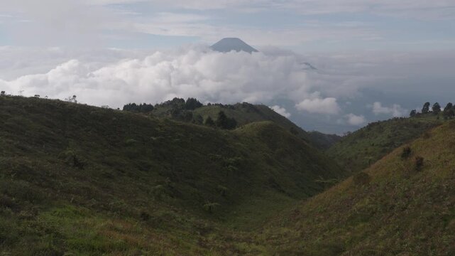 Aerial view of Gunung Prau with a hiker in an orange jacket standing on a grassy ridge overlooking a sea of clouds in Bawang, Central Java, Indonesia.