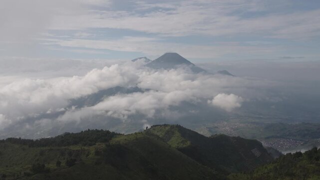 Aerial view of Gunung Prau mountain peaks rising above a thick layer of white clouds with green forested ridges in Bawang, Central Java, Indonesia.