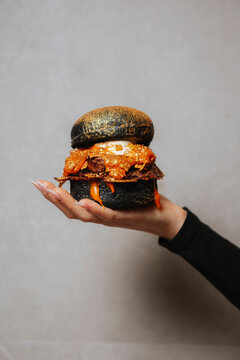 Close-up of a hand presenting a black bun burger with rich toppings in photographic style on a neutral background, concept of gourmet street food