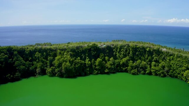 Aerial view of the vibrant green water of a volcanic crater lake surrounded by dense tropical forest and the blue sea in the distance in Ternate, North Maluku, Indonesia.