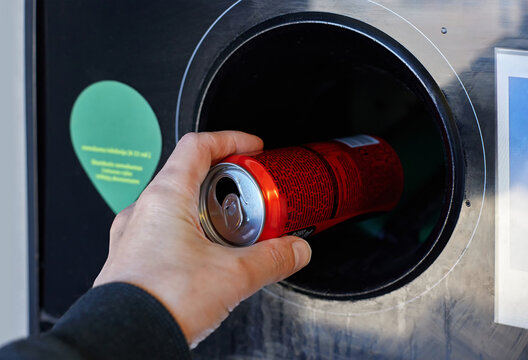 Aluminum can recycling scene showing a hand placing a red beverage container into a circular intake of a reverse vending machine, close-up. An automated deposit-return system. Zero waste lifestyle
