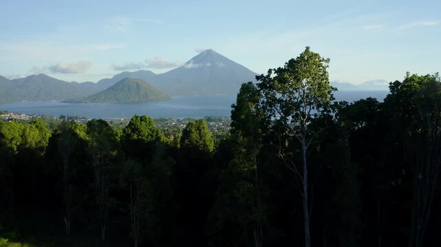 Aerial view of dense tropical trees and distant volcanic peaks across the sea under a clear sky in Ternate, North Maluku, Indonesia.