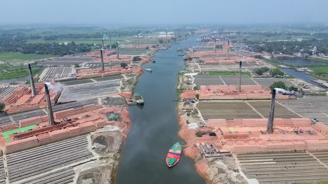 Aerial view of brick kilns with tall chimneys emitting smoke along a river bank with small boats in Narayanganj, Dhaka Division, Bangladesh.
