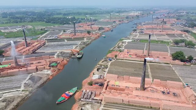 Aerial view of brick kilns with tall chimneys emitting smoke along a river with boats in Narayanganj, Dhaka Division, Bangladesh.