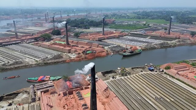 Aerial view of numerous brick kilns with tall chimneys emitting white smoke along a river with cargo boats and stacks of red bricks in Narayanganj, Dhaka Division, Bangladesh.