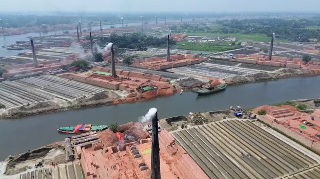 Aerial view of traditional brick kilns with tall chimneys emitting smoke along a river with cargo boats in Narayanganj, Dhaka Division, Bangladesh.
