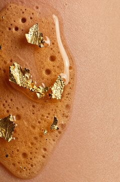 Translucent amber gel with aerated microbubbles clings to human skin, suspended with jagged flakes of brilliant gold leaf that catch the soft overhead light.