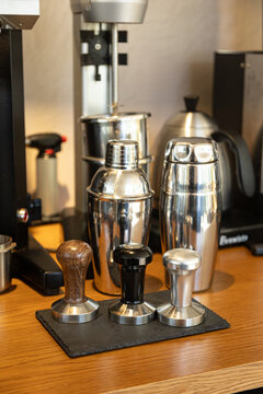 Barista tools and coffee preparation equipment on a wooden counter, including stainless steel milkshake shakers and coffee tampers. Professional coffee setup in a caf&eacute; environment