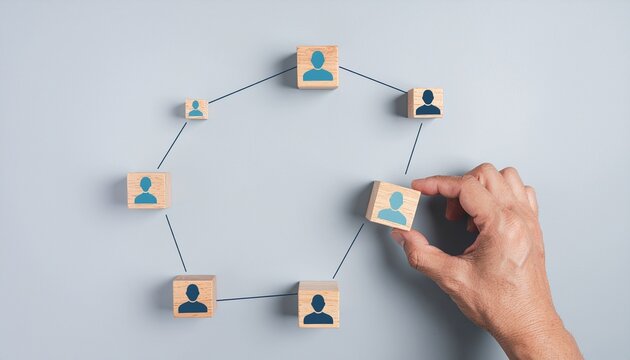 A hand connecting a wooden block with a human icon to a network of similar blocks on a plain gray background, symbolizing social connections or teamwork.