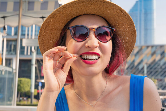 Happy woman wearing sunglasses and a straw hat while using a toothpick, celebrating vacation lifestyle in the urban landscape