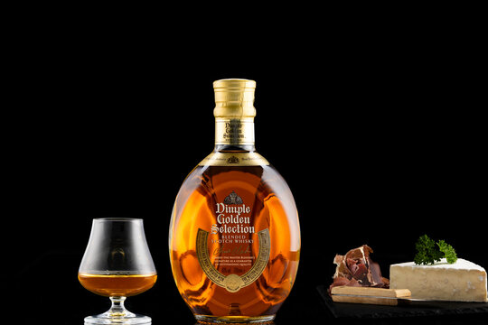 Dimple Golden Selection blended Scotch whisky bottle with tasting glass on black background, styled with cheese and cured meat, illustrative editorial premium spirits concept.