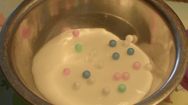 Metal bowl filled with white slime and beads, small hands squishing and mixing tactile foam, playful pastel beads bobbing on surface, messy joyful exploration and learning