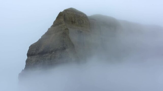 Panning slow motion shot of cliff and rocky headland emerging from dense fog along rugged ocean coastline in overcast morning