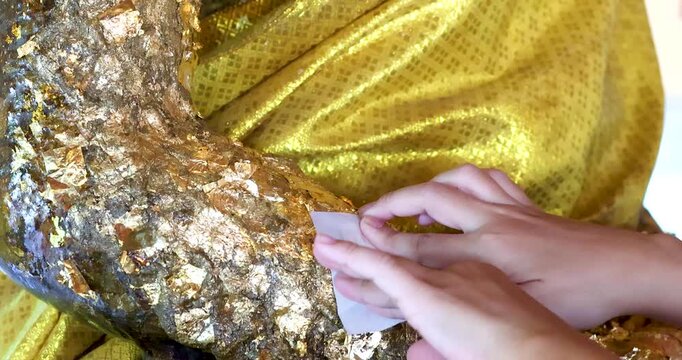Hands applying gold leaf to a Buddha statue symbolizing Buddhist faith, merit making, spirituality, Thai culture, meditation, mindfulness, and religious tradition, close-up