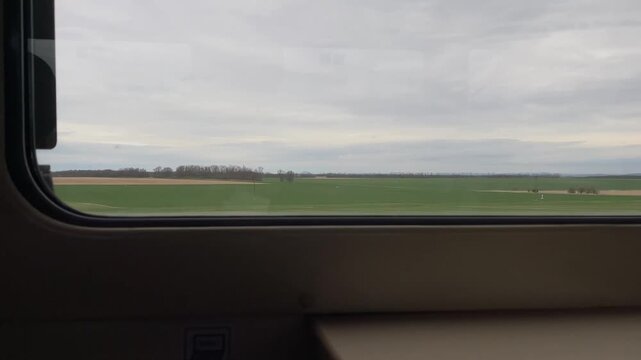 Train window view across green fields, flat farmland with pond and distant tree line, overcast sky and gentle motion, calm commuter mood, photographer capturing passing panorama