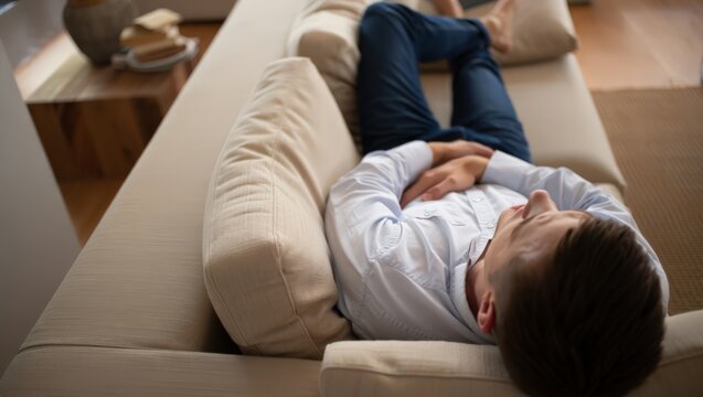 A man lies on a couch holding his stomach, looking unwell in a modern living room with a wooden coffee table and beige rug.