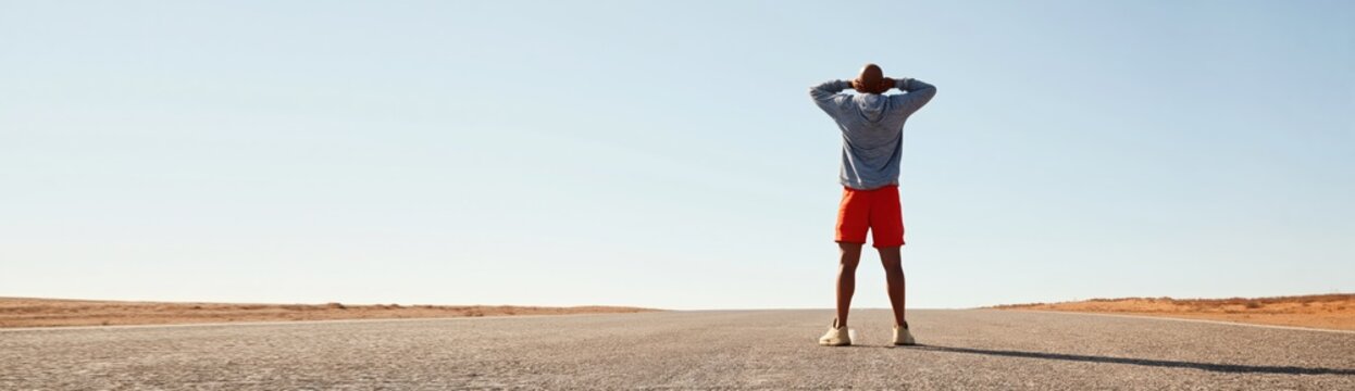 Person standing with hands behind head on sunlit road in open flat landscape