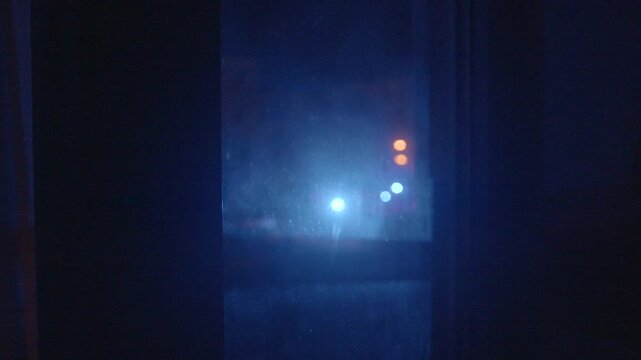 Night scene emergency vehicle lights flashing outside window, red and blue beams, hazy reflection on glass, urgent atmosphere with distant siren impression and street illumination