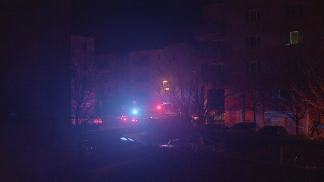Night street illuminated by flashing lights, emergency vehicles respond outside apartment building, red and blue strobes cast silhouettes and shadows, siren wail and urgent