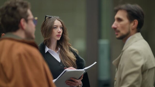 Three business people are having a discussion outdoors in an urban environment. A young professional woman is seen taking notes during their city meeting.