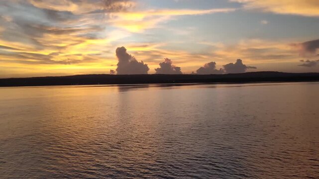 A golden sunset glows over a calm sea, casting warm light across the water. Silhouetted hills and unique, upright clouds sit on the horizon under a soft, painted sky.