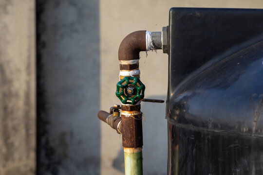 Close-up of a green gate valve attached to a rusty iron pipe on a black plastic water storage tank. Ideal for plumbing services, water management, and home maintenance themes.