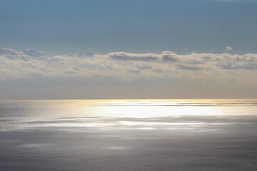 Obraz premium A tranquil seascape where sunlight pierces through clouds and glistens across the calm water surface. The soft golden reflections and layered clouds create a peaceful, atmospheric horizon scene.