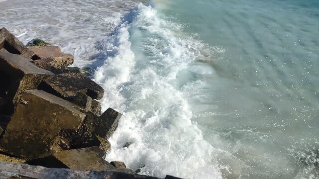 Waves crashing against dark tetrapod rocks on a sandy beach. The turquoise water creates white sea spray and foam under bright sunlight. Clear and refreshing coastal scenery.