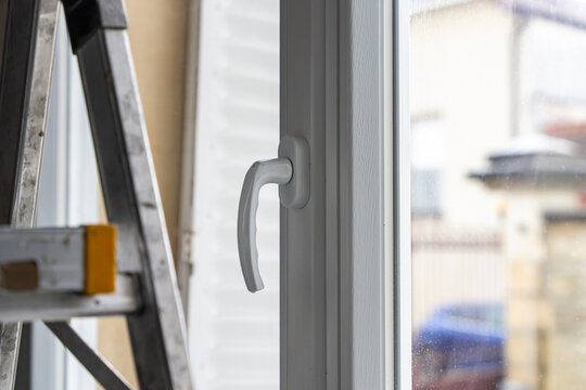 Gros plan sur une poign&eacute;e de fen&ecirc;tre moderne en PVC blanc, symbole d'une installation ou d'une r&eacute;novation r&eacute;ussie, avec un escabeau &agrave; proximit&eacute; &eacute;voquant les derni&egrave;res finitions int&eacute;rieures.