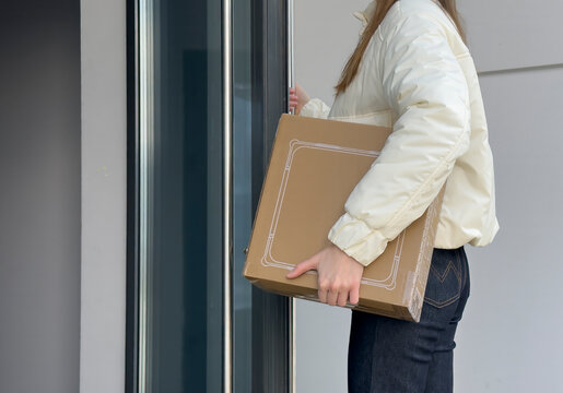 Woman carries a cardboard box at an apartment door. She opens the glass entrance during a home delivery or move. Residential relocation and parcel service concept..